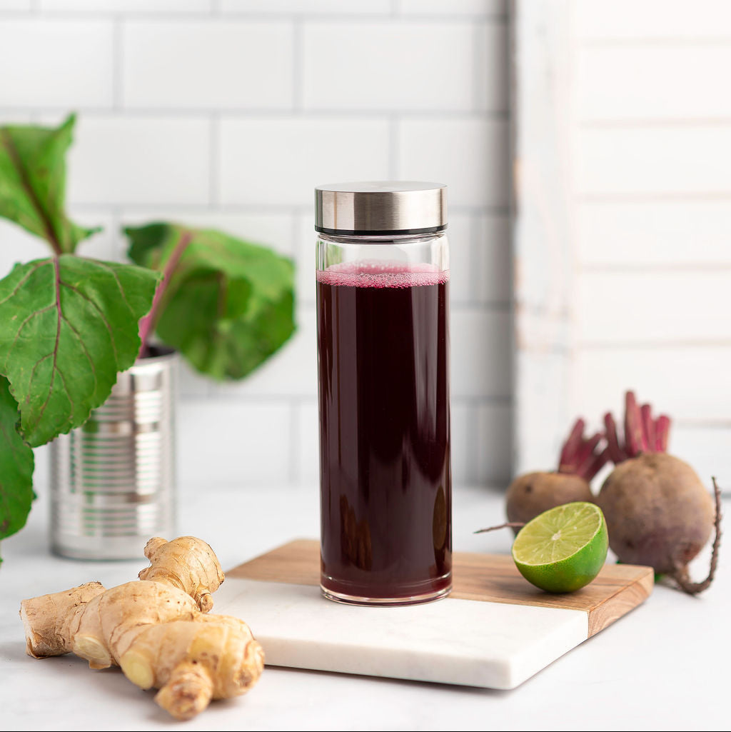 Glass of dark juice with beetroot, ginger, and lime on a cutting board against a white tiled wall.