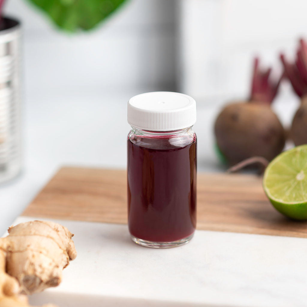 Jar of beet juice shots on a cutting board with ginger and lime and raw beets in the background