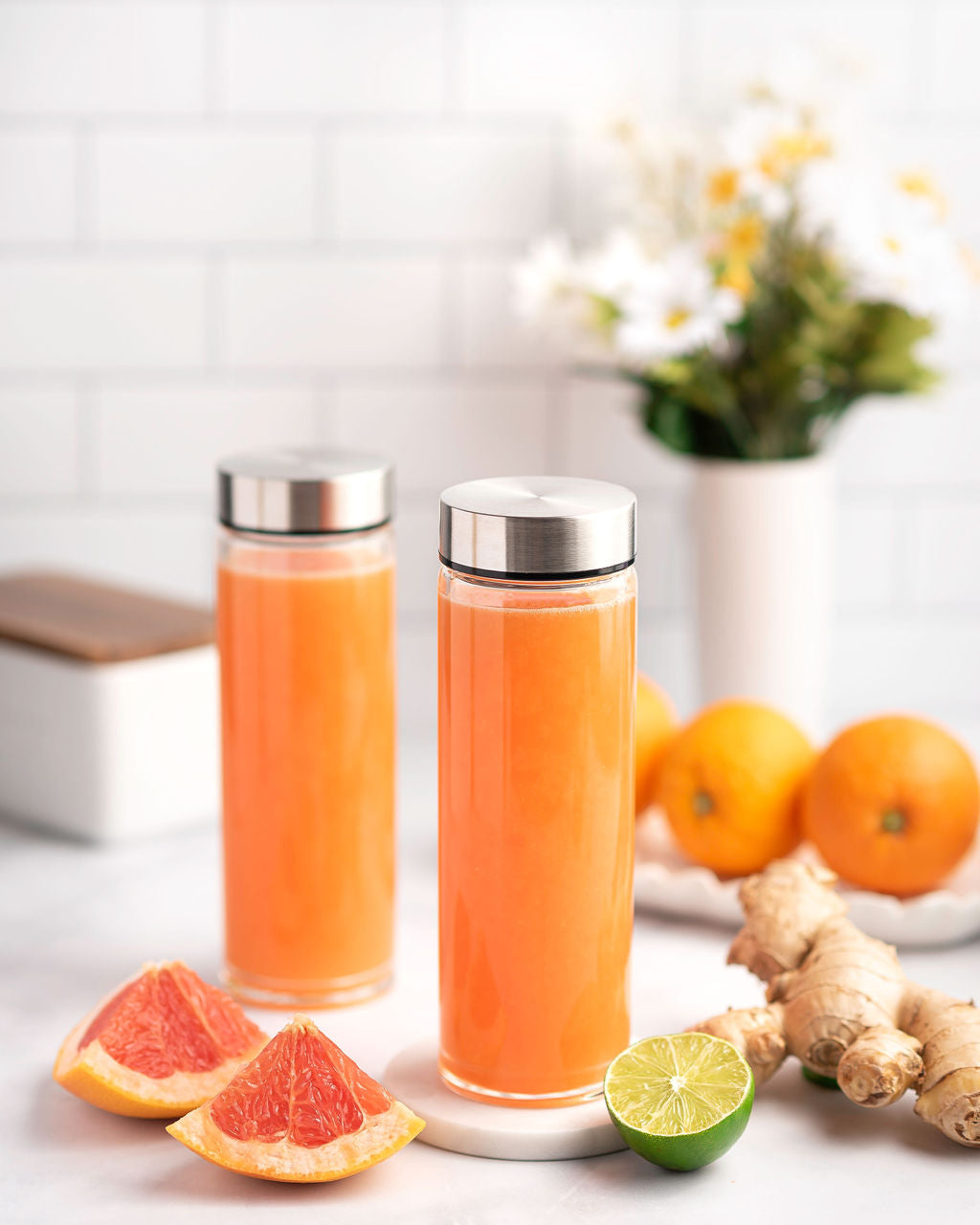 Two glass bottles with silver lids filled with orange juice, surrounded by citrus fruits and ginger on a light background.