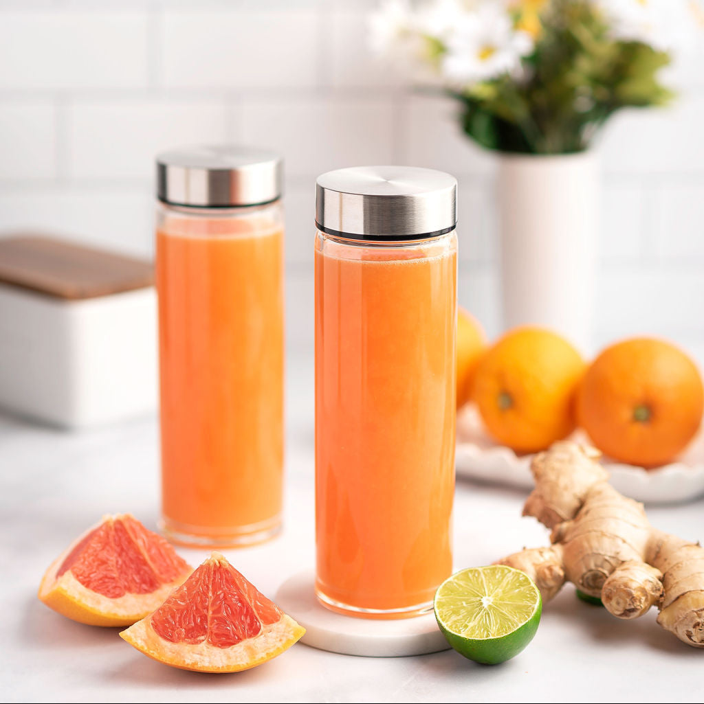 Two glass bottles with silver lids filled with orange juice, surrounded by citrus fruits and ginger on a light background.