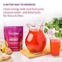 PuriVibe Clean Energy Yerba Mate Energy drink mix in a purple pouch, shown next to a glass pitcher filled with red juice and a glass of the same drink on a wooden board, with fruit pieces and a plant in the background.