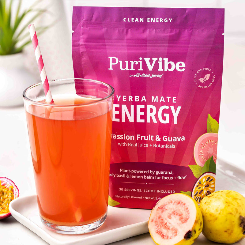 A stand-up pouch of PuriVibe Yerba Mate Energy Passion Fruit & Guava drink mix stands behind a clear glass filled with pink-orange beverage and a pink-and-white striped straw. The scene includes sliced passion fruit and a whole guava in a bright, clean setting.